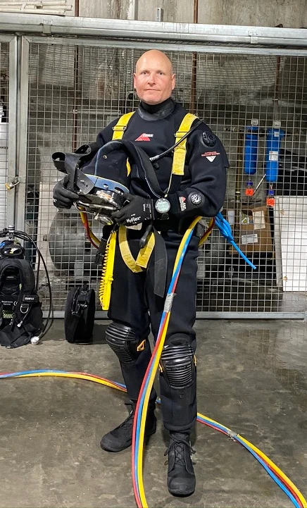 Israel Hannant from Fire Tanks Australia wearing diving equipment and holding a diving helmet, with hoses in front of a metal gate.