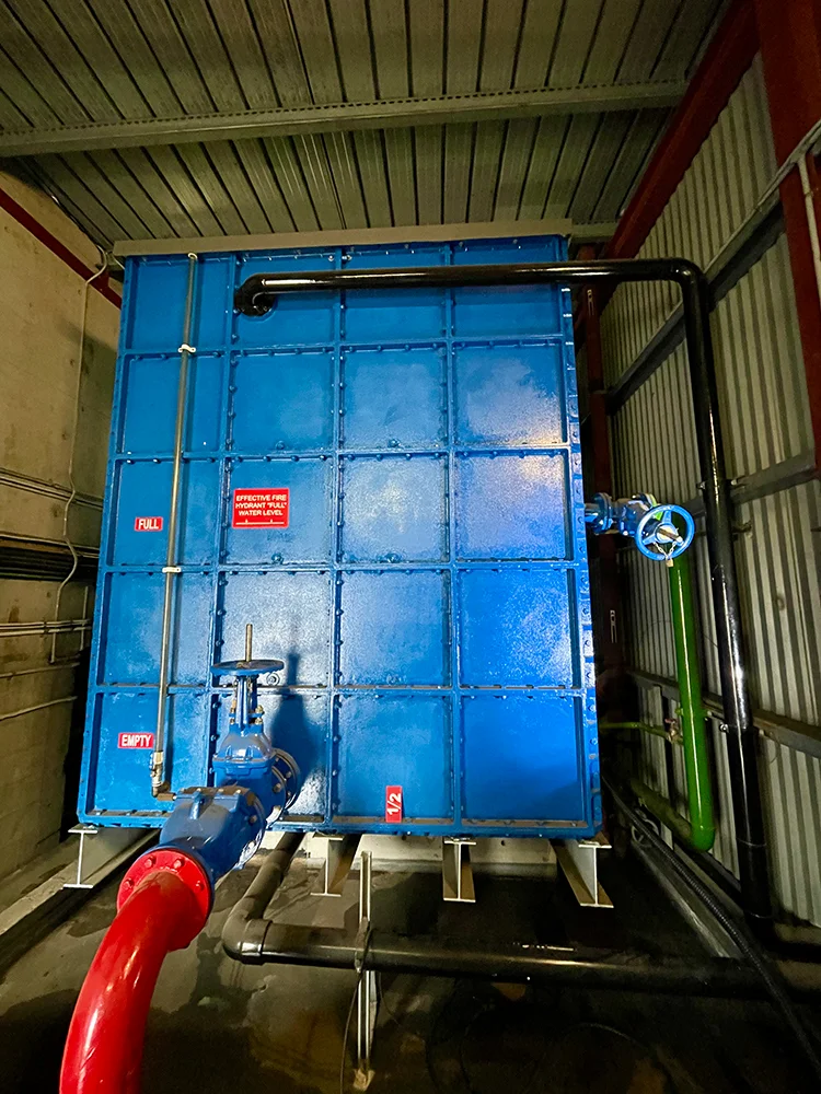 Blue sectional water tank with labeled water level indicators, connected pipes, and safety signage inside a fire protection facility.
