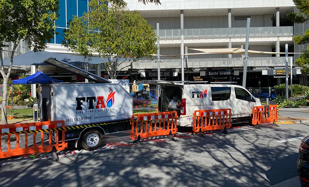 Fire Tanks Australia van and trailer parked on-site, surrounded by orange safety barriers, with a commercial building in the background.